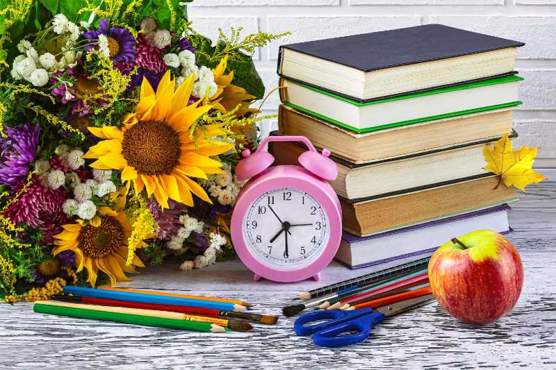 A pile of books a pink alarm clock stationery an apple and autumn flowers