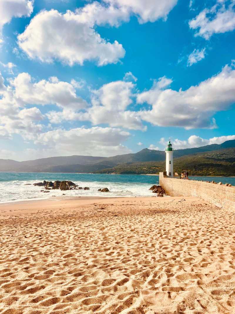 A tranquil beach scene with a lighthouse and mountains under a blue sky