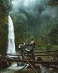 two person carrying black inflatable pool float on brown wooden bridge near waterfalls - jigsaw puzzle