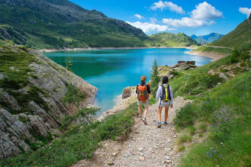 two friends during a hike in the mountains walking on path near an alpine lake