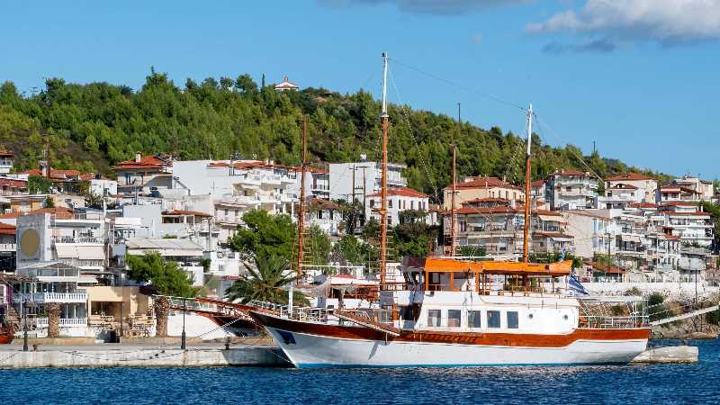 Two moored sailboats near a pier in neos marmaras buildings located on a hill with multiple greenery